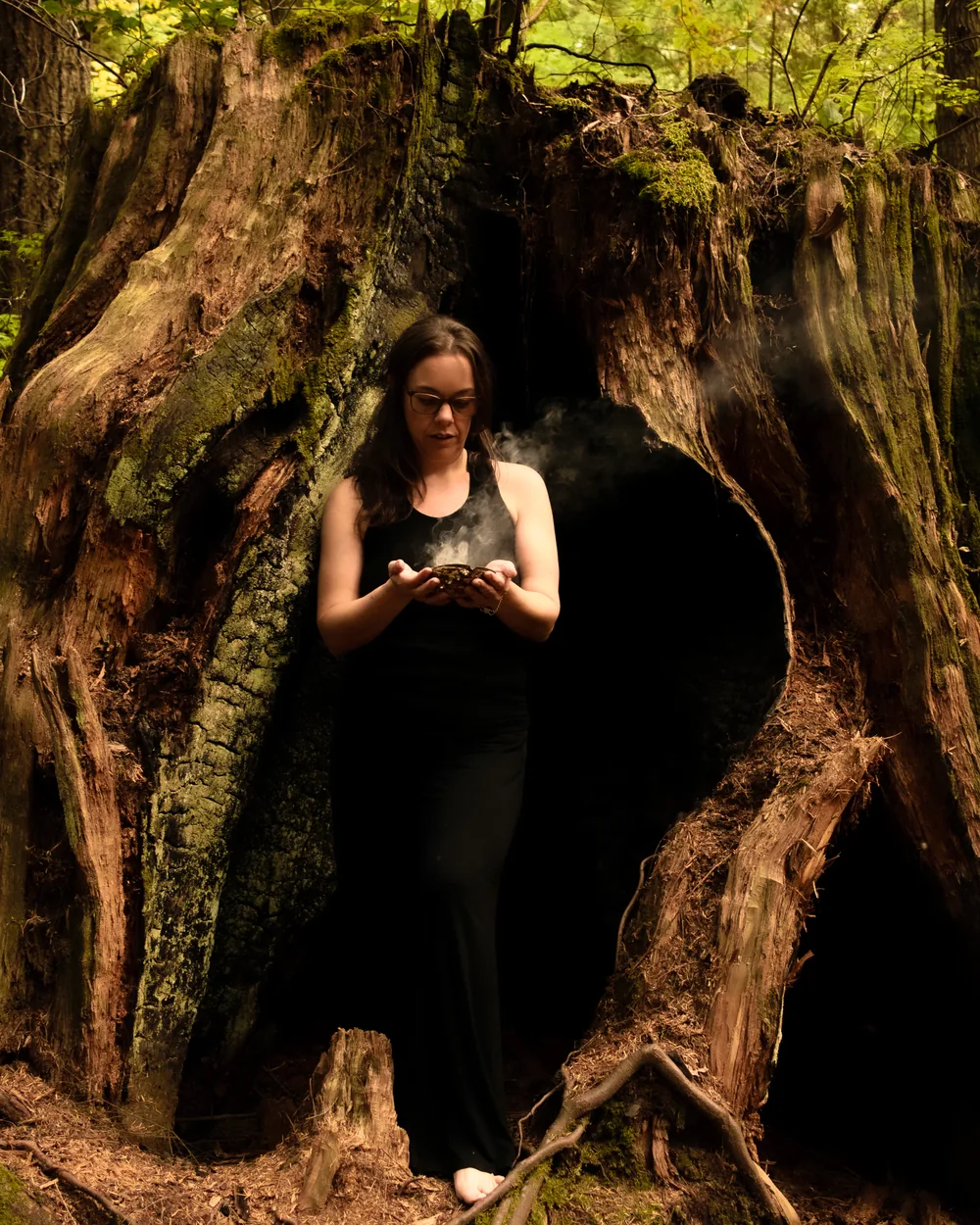 Fernanda standing barefoot inside the hollow of an old cedar, holding a smoking shell